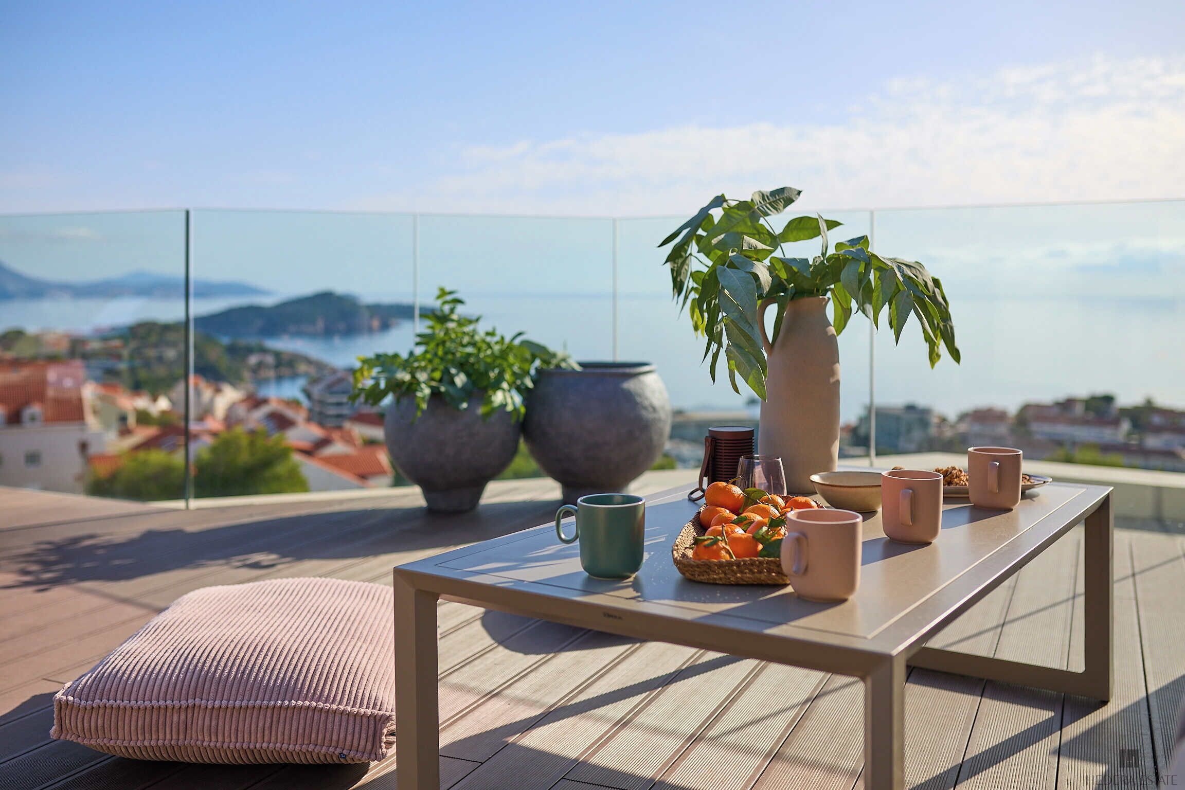 A terrace with a table set for two, a potted plant, and a view of the sea and hills.