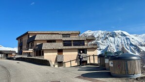 A multi-story building with a ski lift in the background, a person standing in front, and a mountain range. - LE BELLEVUE (Orcières)