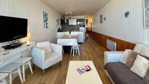 A modern living room with a flat-screen TV, white sofas, a dining table, and a kitchen in the background. - LE BELLEVUE (Orcières)