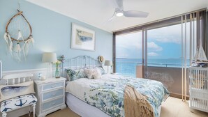Une chambre avec une grande fenêtre donnant sur la mer, un lit avec une literie à motifs floraux, une table de chevet, une commode et un attrape-rêves accroché au mur.