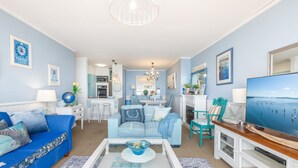 Un salon avec un canapé bleu, une table basse et un téléviseur. Au fond, on aperçoit une cuisine avec un coin repas.