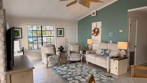 Un salon doté d’un ventilateur de plafond, d’un canapé, de deux fauteuils, d’une table basse, d’un téléviseur et d’une fenêtre donnant sur une piscine.