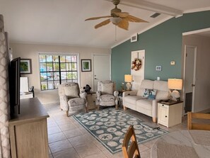 A living room with a ceiling fan, a sofa, two armchairs, a coffee table, a TV, and a window with a view of a pool.