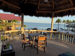 A covered outdoor seating area with a table and chairs, overlooking the sea and a dock with boats.