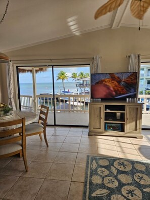 A living room with a TV, a dining table, chairs, a view of the ocean, and a balcony with a railing.