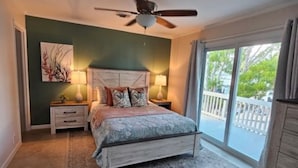 Une chambre à coucher comprenant un lit, deux tables de chevet avec des lampes, un ventilateur de plafond, une grande fenêtre avec vue et un balcon.