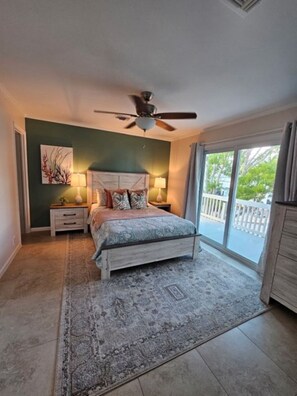 A bedroom with a bed, two bedside tables with lamps, a ceiling fan, a large window with a view, and a balcony.