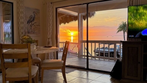 Un coin repas avec une table et des chaises, un téléviseur posé sur un meuble, et une vue sur le coucher de soleil à travers une porte vitrée.