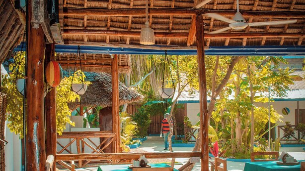 An outdoor dining area with wooden tables and chairs, a thatched roof, and hanging lights. - Tuna Nungwi Beach Bungalow (Nungwi)