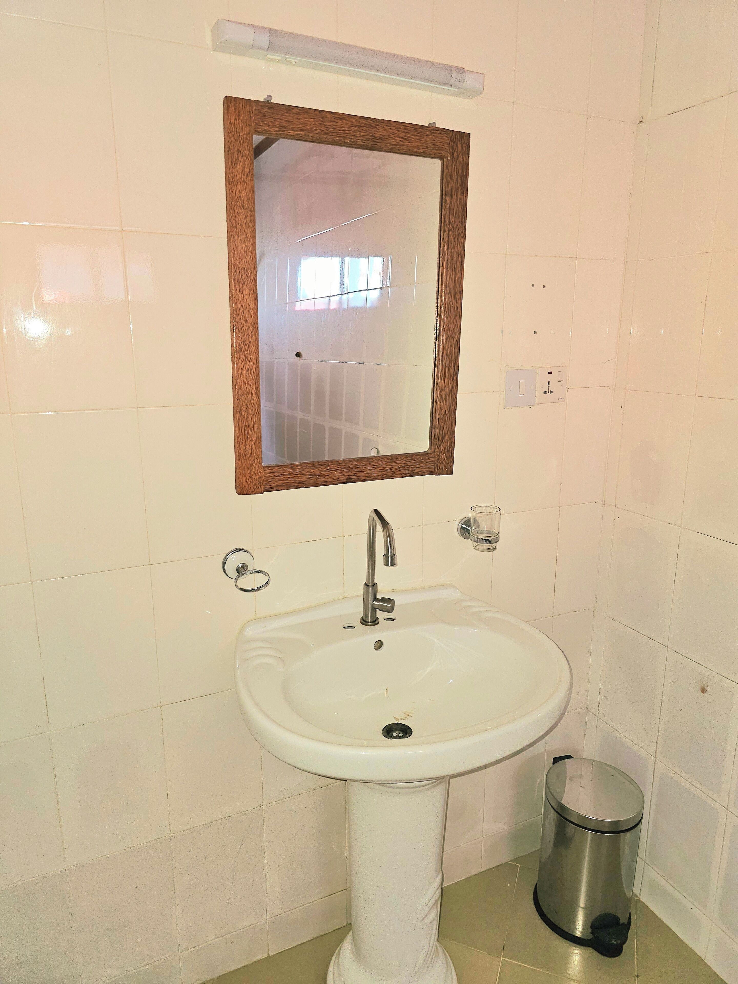 Un baño con un lavamanos blanco, un espejo con marco de madera, un grifo, un cesto de basura y paredes de azulejos.