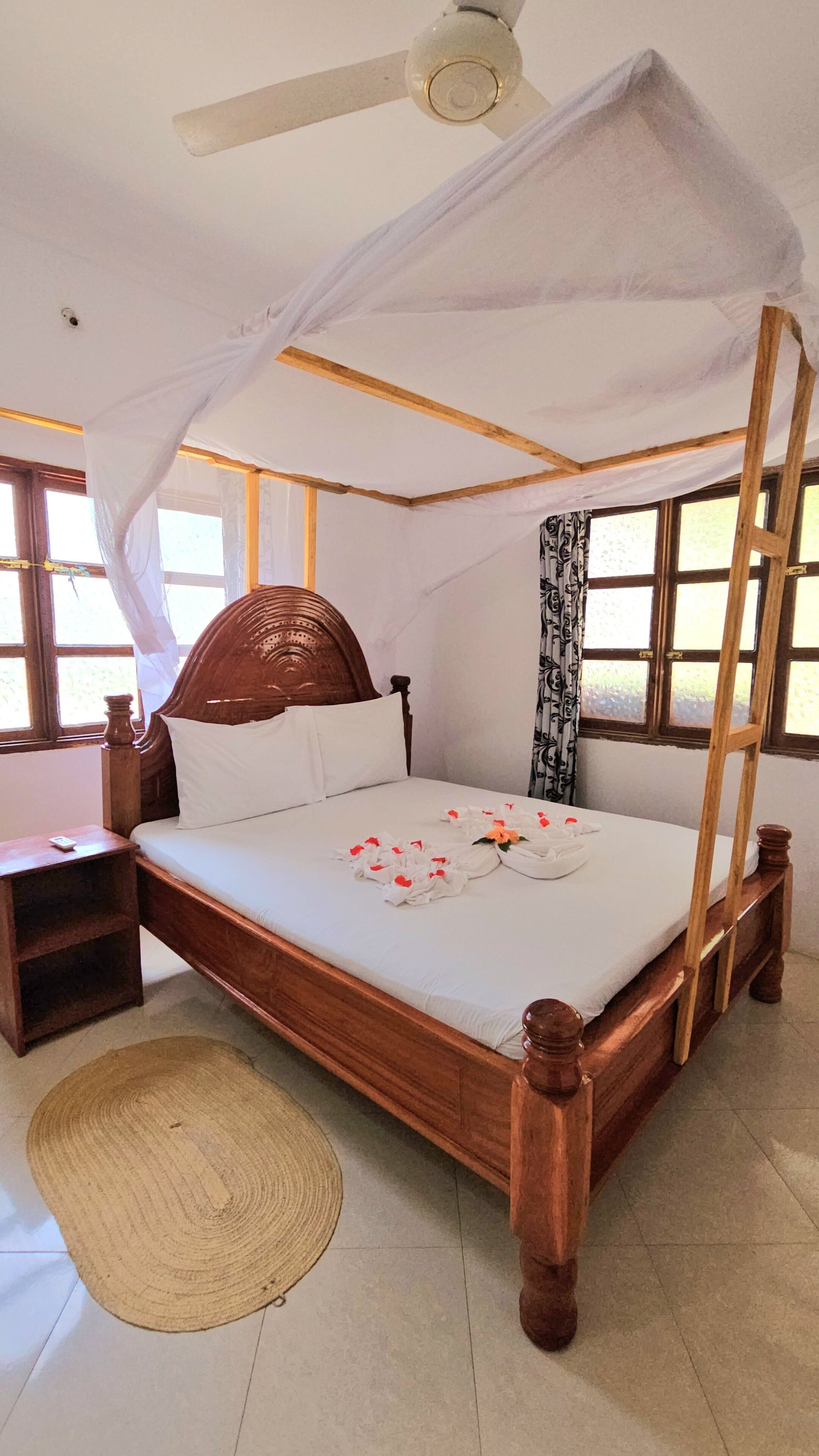 Un lit en bois avec un baldaquin, une table de chevet, un tapis tissé et un ventilateur de plafond.