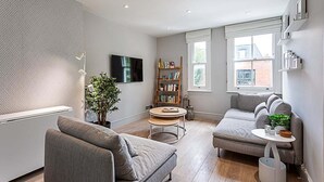 Uma sala de estar moderna com um sofá cinza, uma mesa de centro redonda, uma estante e uma TV fixada na parede.