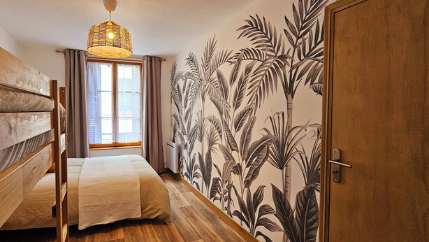 Ein schmales Zimmer mit einem Bett, einem Holzschrank, einem Fenster mit Vorhängen und einer Wand mit einem Wandbild von tropischer Vegetation.