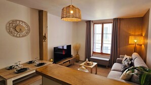 Ein modernes Wohnzimmer mit einem Flachbildfernseher, einem Holzesstisch gedeckt mit Tellern und Gläsern, einer Couch mit Kissen, einem großen Fenster mit Vorhängen und einer Pendelleuchte.