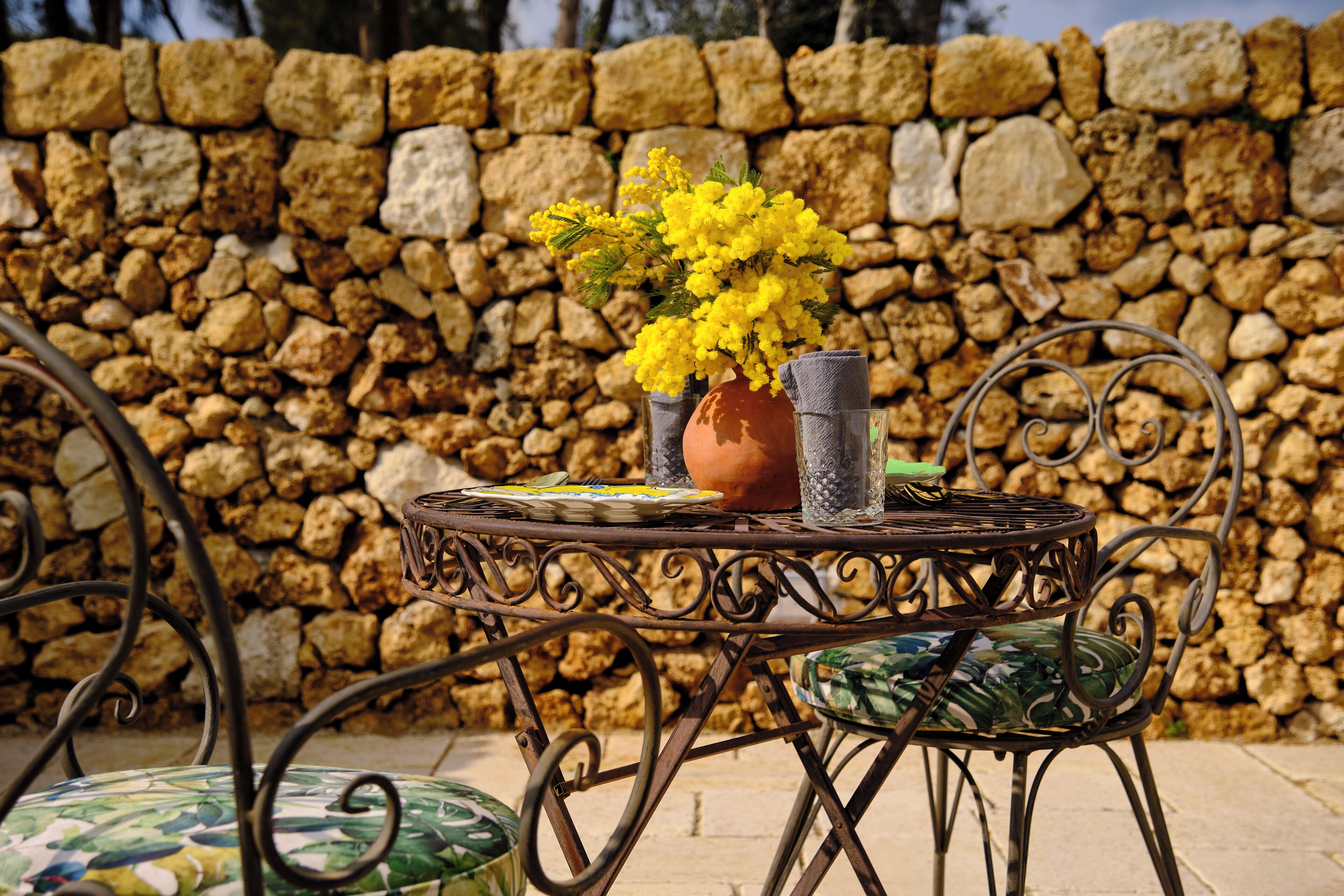 A stone wall, a metal table with chairs, and a vase with yellow flowers.