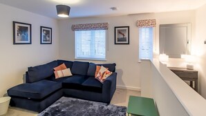 A living room with a dark blue sectional sofa, a small rug, and a green stool. There are framed pictures on the wall, a window with blinds, and a staircase with a mirror. - Charming Coastal Retreat – 1-Bedroom Flat in Blyth - Pass the Keys (Blyth)