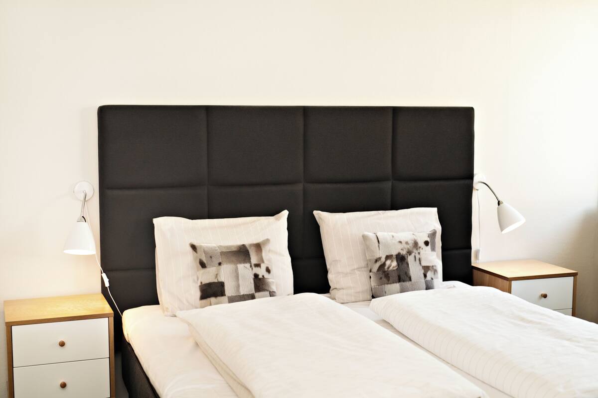 A neatly made bed with a black headboard, two bedside tables with lamps, and a small wooden nightstand with a drawer.