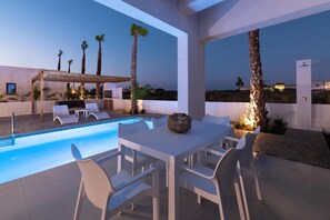A modern outdoor pool area with a dining table and chairs, surrounded by palm trees and a clear view of the landscape.