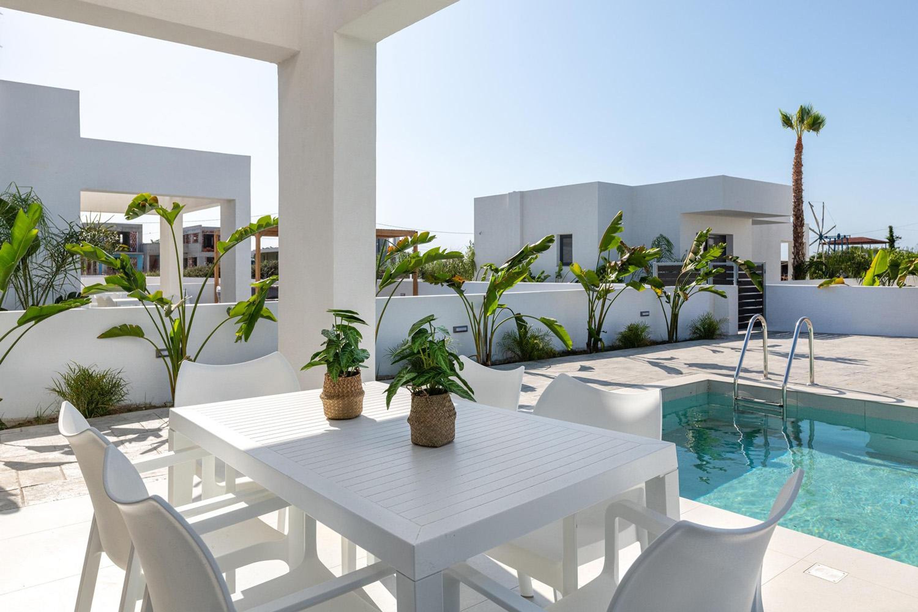 A poolside area with a white table and chairs, surrounded by potted plants and a clear pool.