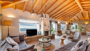 A spacious living room with a fireplace, wooden ceiling beams, and a large TV. - Villa Lithos in Paxi (Paxos)