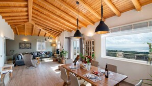 A spacious dining area with a wooden table, chairs, and pendant lights. A large window offers a view of the outdoors. - Villa Lithos in Paxi (Paxos)