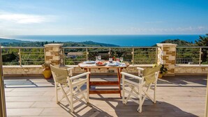 A terrace with a table set for two, overlooking a scenic view of the sea and hills. - Villa Lithos in Paxi (Paxos)