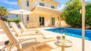 Un área junto a la piscina con sillones de descanso y una mesa, una piscina y un edificio con tejado de baldosas.