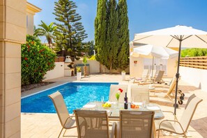 A poolside area with a table set for dining, lounge chairs, and a large umbrella. - Villa Clara in Paralimni (Paralimni)