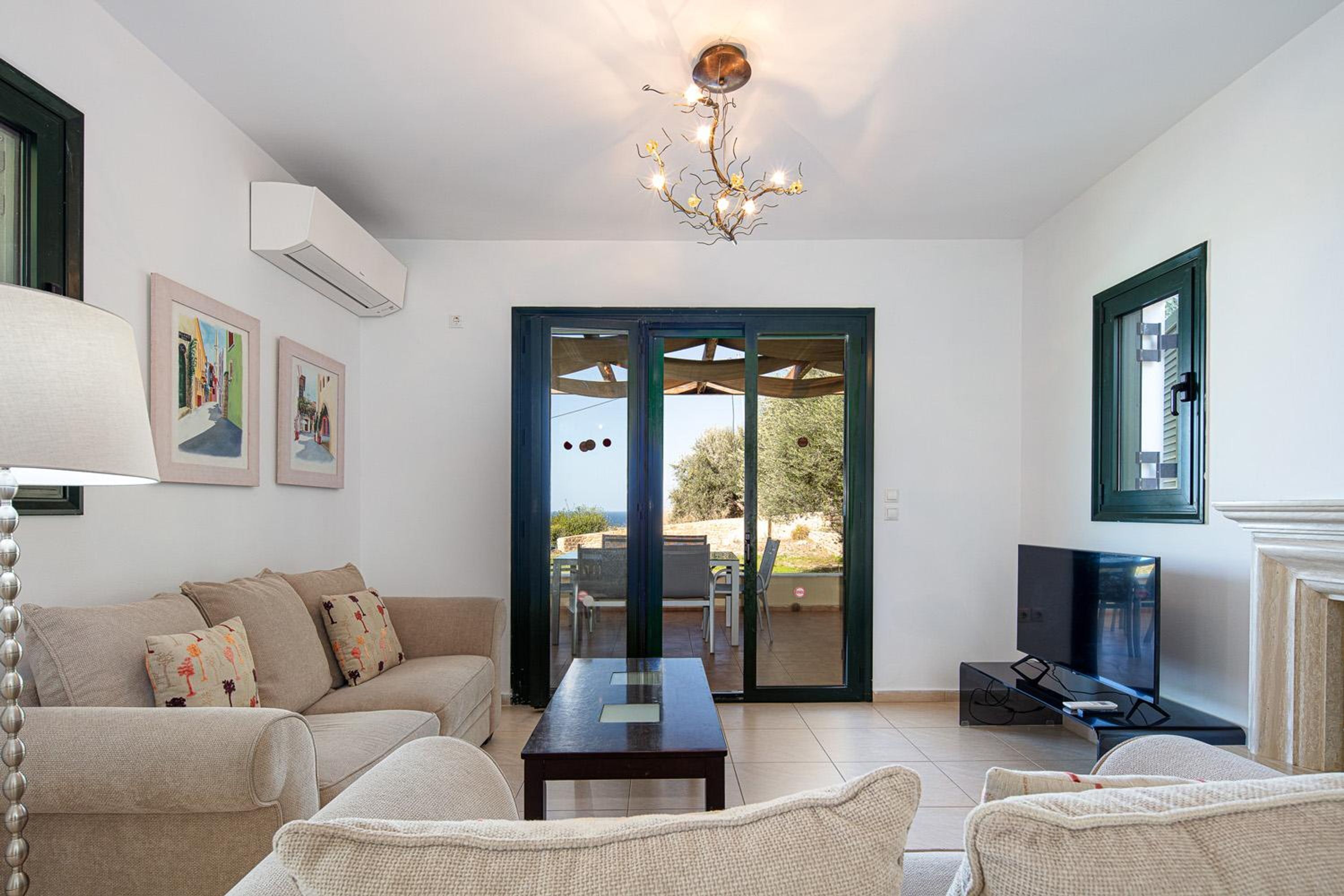 A living room with a sofa, a coffee table, a TV, and a chandelier. There is a door leading to an outdoor area with a view of the sky.