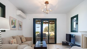 A living room with a sofa, a coffee table, a TV, and a chandelier. There is a door leading to an outdoor area with a view of the sky. - Villa Eleni in Pigianos Kampos (Rethymno)