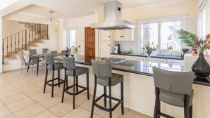 A spacious kitchen with a central island, a dining area with bar stools, a staircase, and a large window providing natural light. - Villa Claudia in Paralimni (Paralimni)