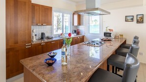 A modern kitchen with wooden cabinets, a granite countertop, and a central island with bar stools. - Villa Koula in Paralimni (Paralimni)