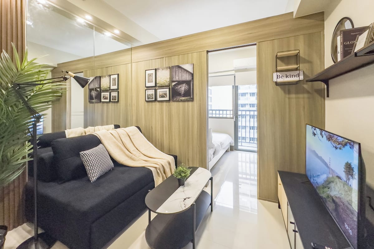 Un salon moderne avec un canapé, une table basse et un téléviseur. On y trouve une plante, des tableaux encadrés au mur et une fenêtre donnant sur des immeubles.