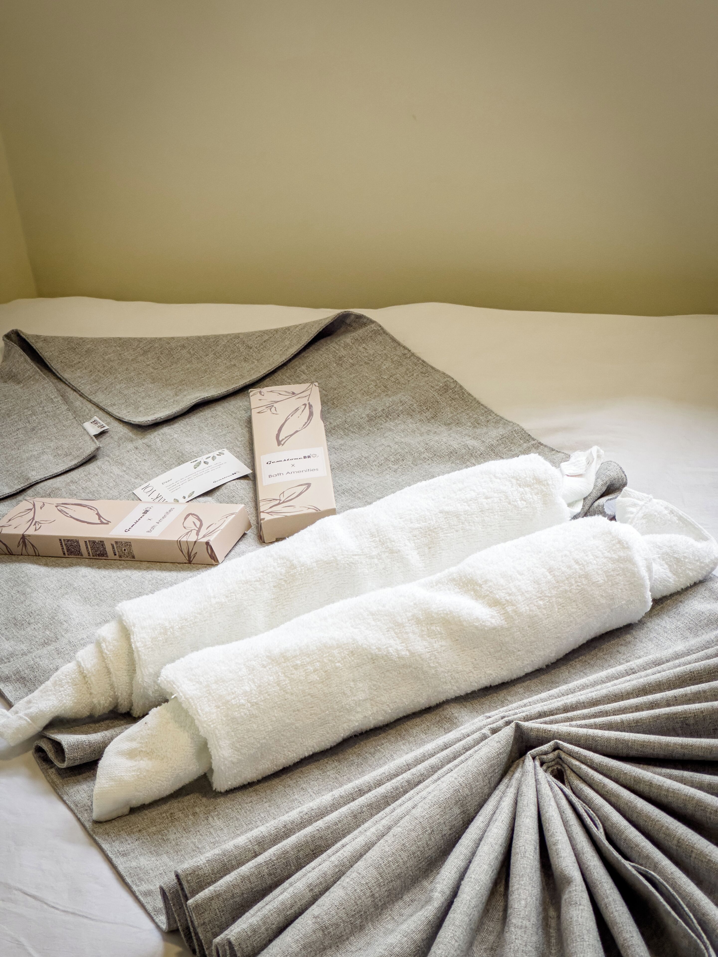 Ein ordentlich bezogenes Bett mit einem gefalteten grauen Handtuch und einem weißen Handtuch, sowie zwei Boxen mit den Aufschriften 'Gemstone BR' und 'Both Amenities'.