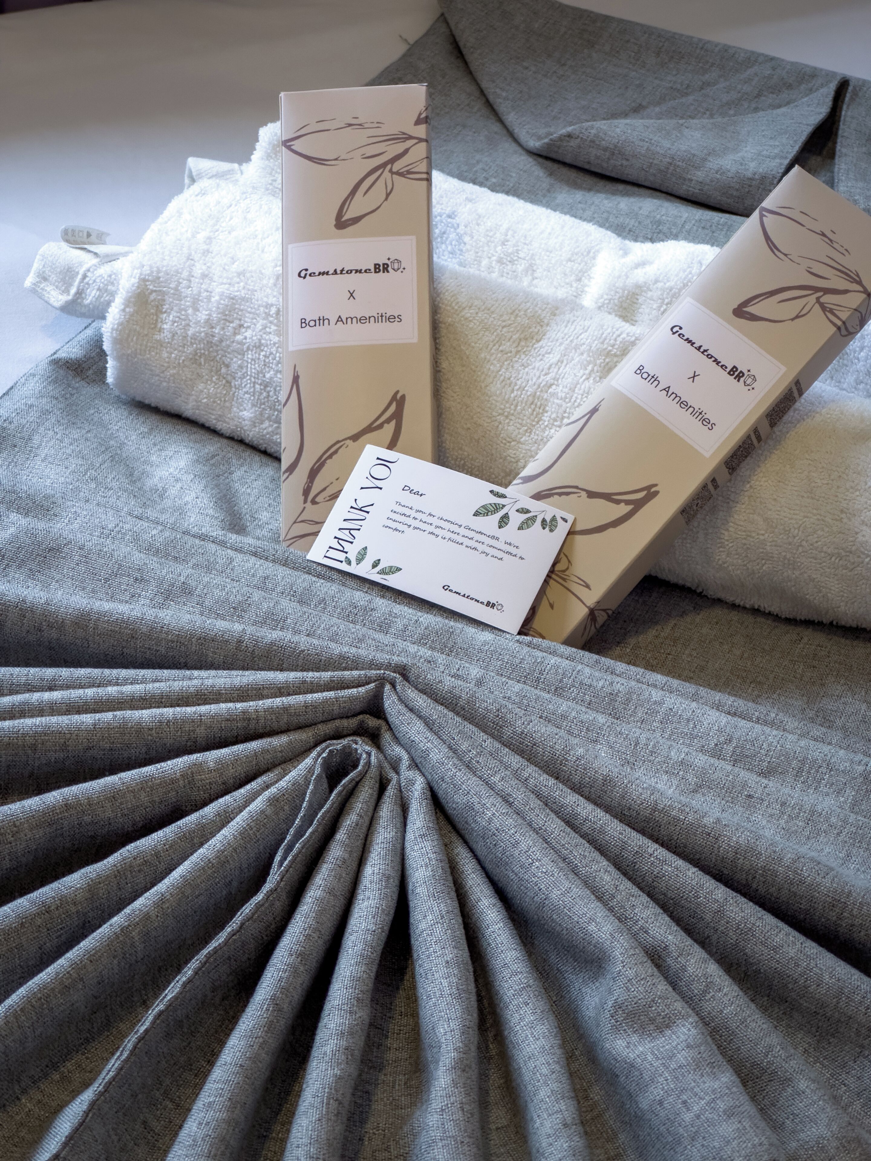A neatly folded grey bath mat with a leaf pattern, placed on a bed with a white towel and a box labeled "GemstoneBR Bath Amenities".