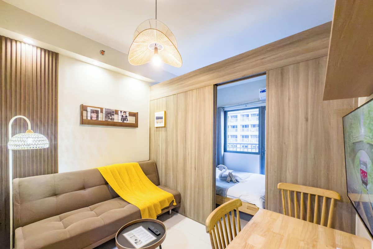 Une chambre d’hôtel moderne avec un canapé, une petite table, une chaise en bois, un lit donnant sur une fenêtre et un luminaire suspendu.