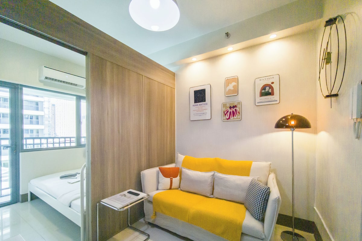 Un salon moderne avec un canapé, une table d’appoint et une lampe sur pied. Des tableaux encadrés ornent le mur, et un appareil de climatisation est fixé au mur.