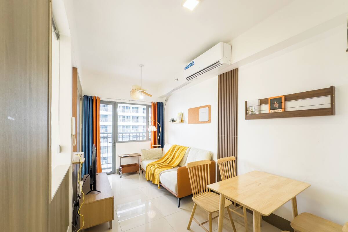 Une chambre d’hôtel moderne avec un canapé, une table à manger en bois, une petite table d’appoint et une chaise. On y trouve une fenêtre avec des rideaux, un appareil de climatisation et une étagère fixée au mur, sur laquelle est posé un cadre photo.
