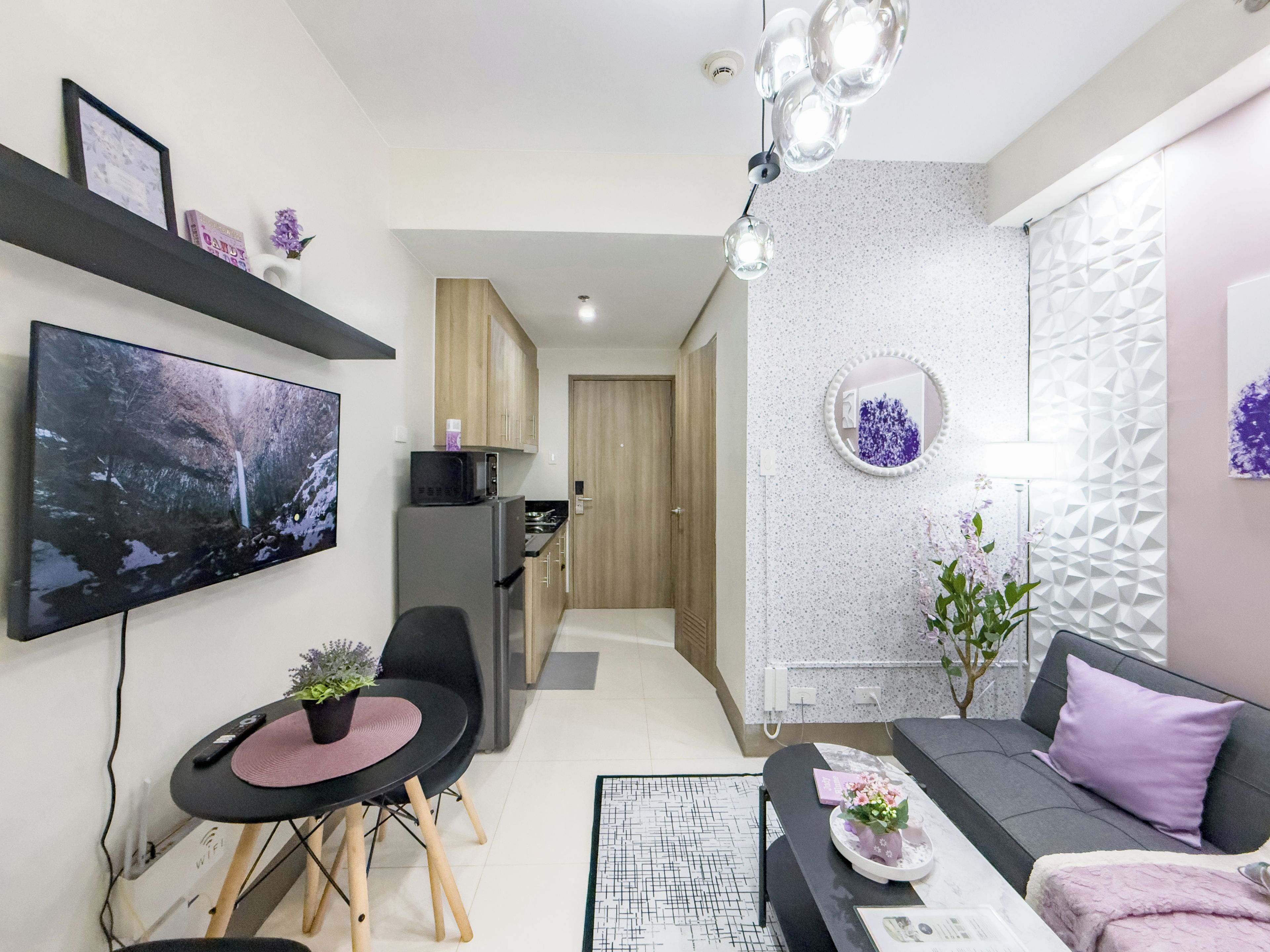 A modern living room with a flat-screen TV, a sofa, a dining table with chairs, and a kitchen area in the background.
