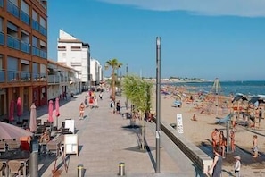 A beachside promenade with a row of buildings on one side, a sandy beach with umbrellas and people on the other, and a clear blue sky.