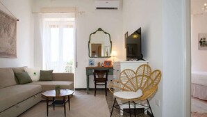 Una sala de estar con un sofá, una mesita, un sillón de mimbre, un escritorio con una silla, un televisor y un espejo.