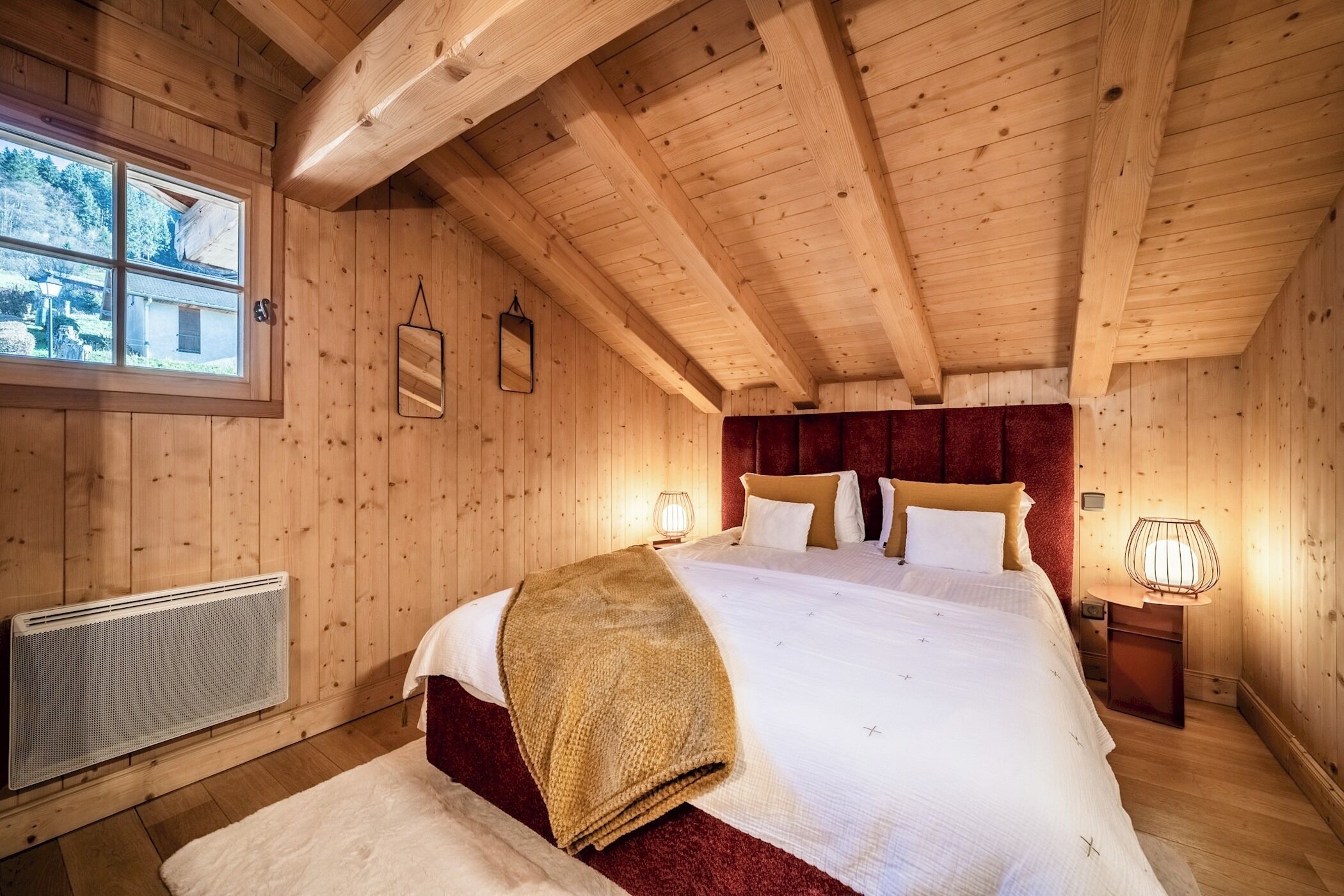 A wooden cabin bedroom with a large bed, a radiator, and a window with a view of the outdoors.