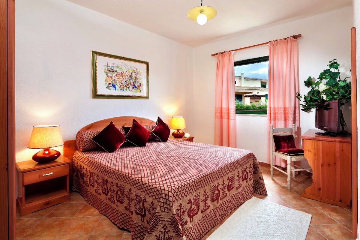 Una camera da letto con un letto, due lampade da comodino, un tavolino, una sedia, un armadio in legno, una pianta in vaso e una finestra con tende.
