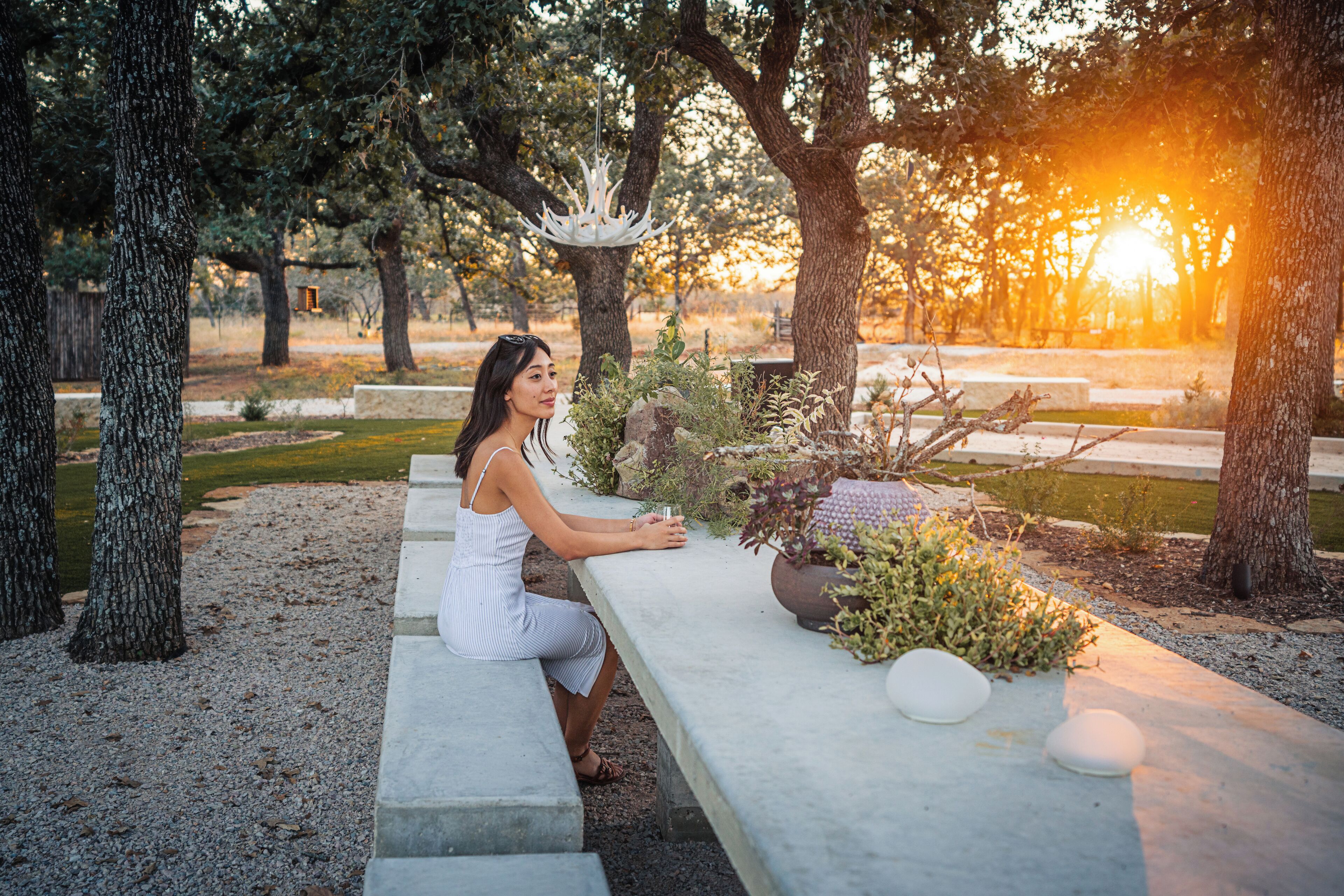 A woman sitting at a concrete bench in a park-like setting with trees and a decorative light fixture.