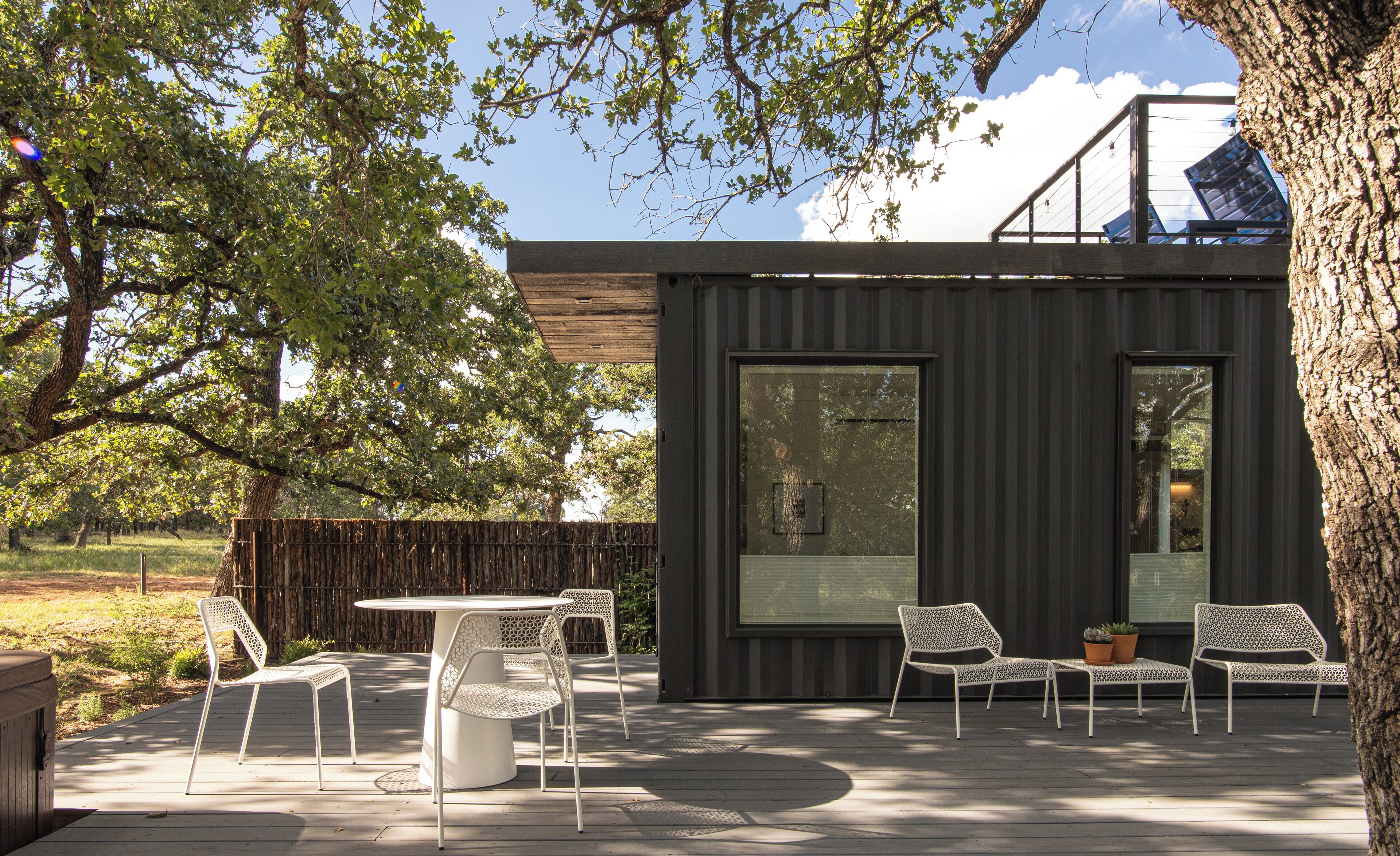 Un patio exterior moderno con una mesa redonda y sillas blancas, una valla de madera y un árbol grande.