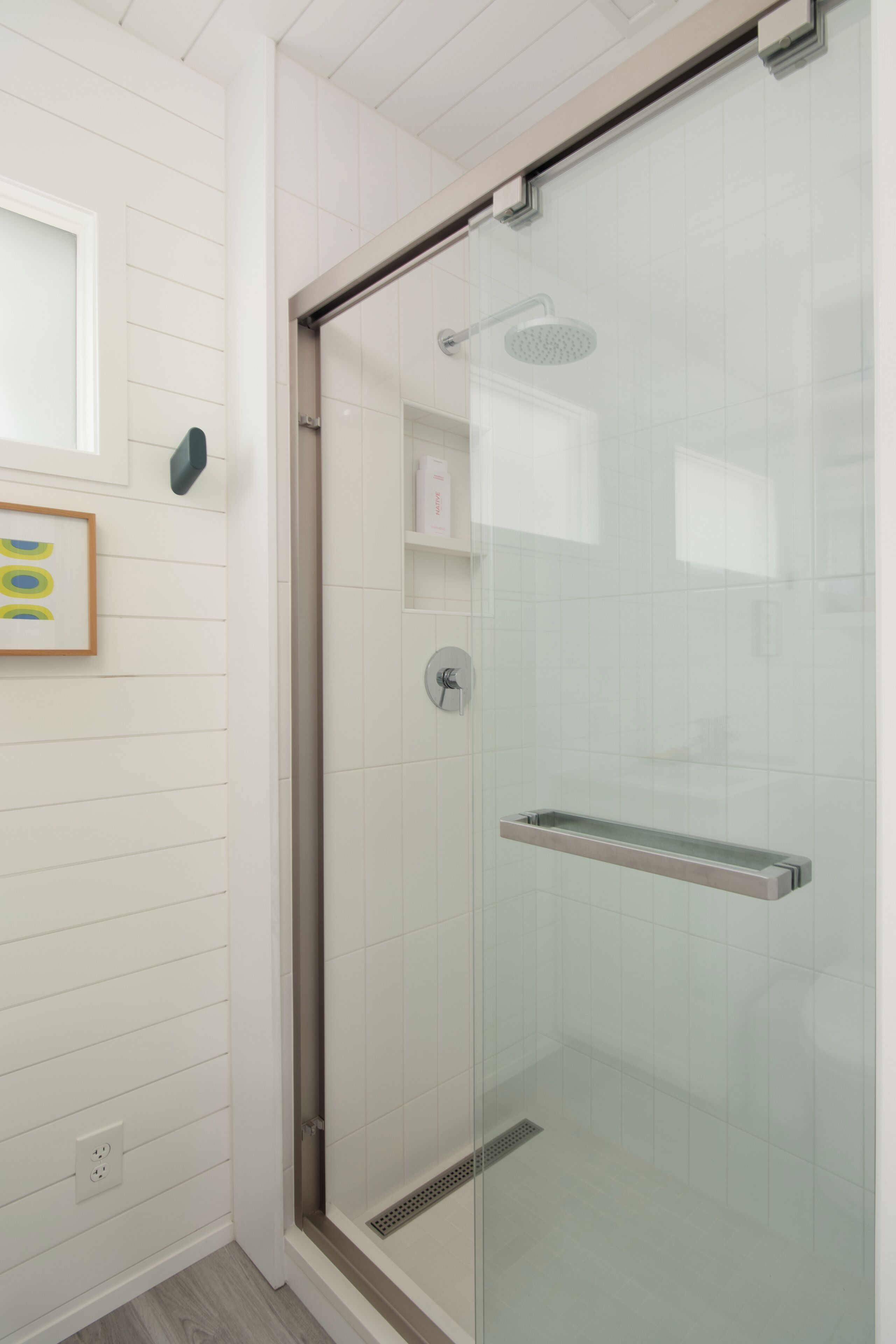 A glass-enclosed shower with a handheld showerhead, a glass shelf, and a tiled wall.