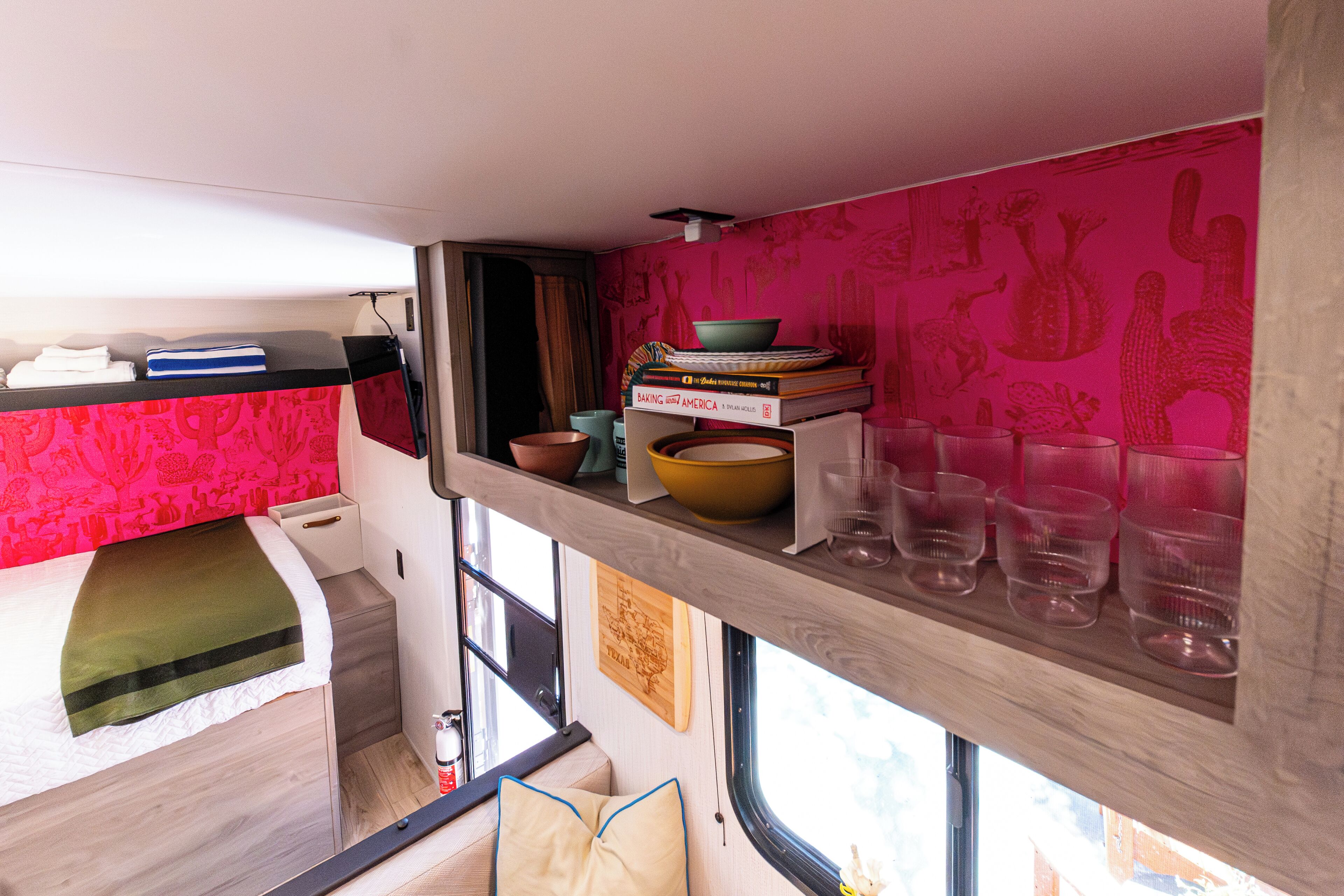 A compact kitchen with a bed adjacent to it, featuring a small shelf with kitchenware and a window providing a view outside.