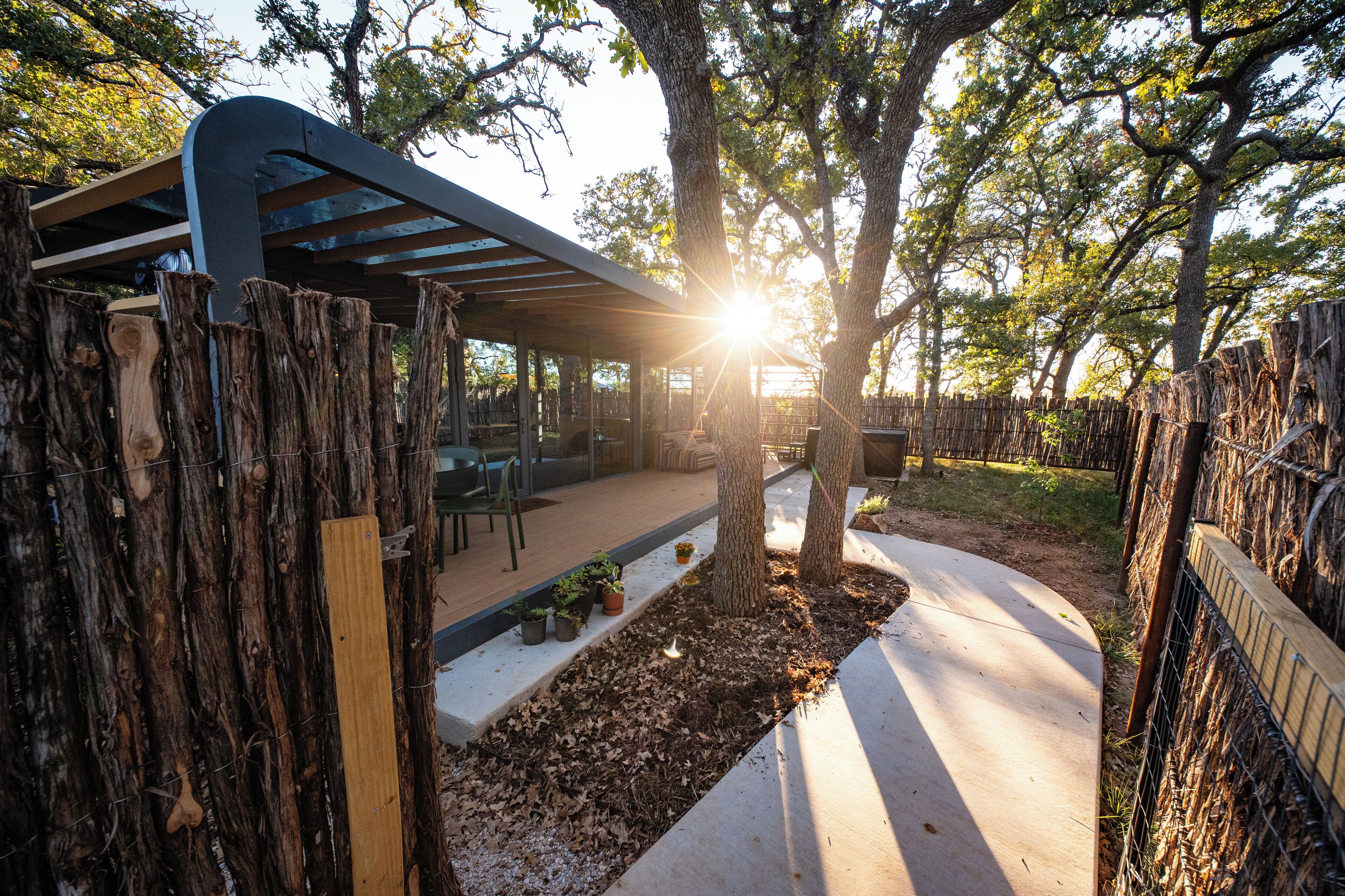 Un espace extérieur aménagé avec une pergola et des sièges, entouré d’arbres et bordé d’un sentier.