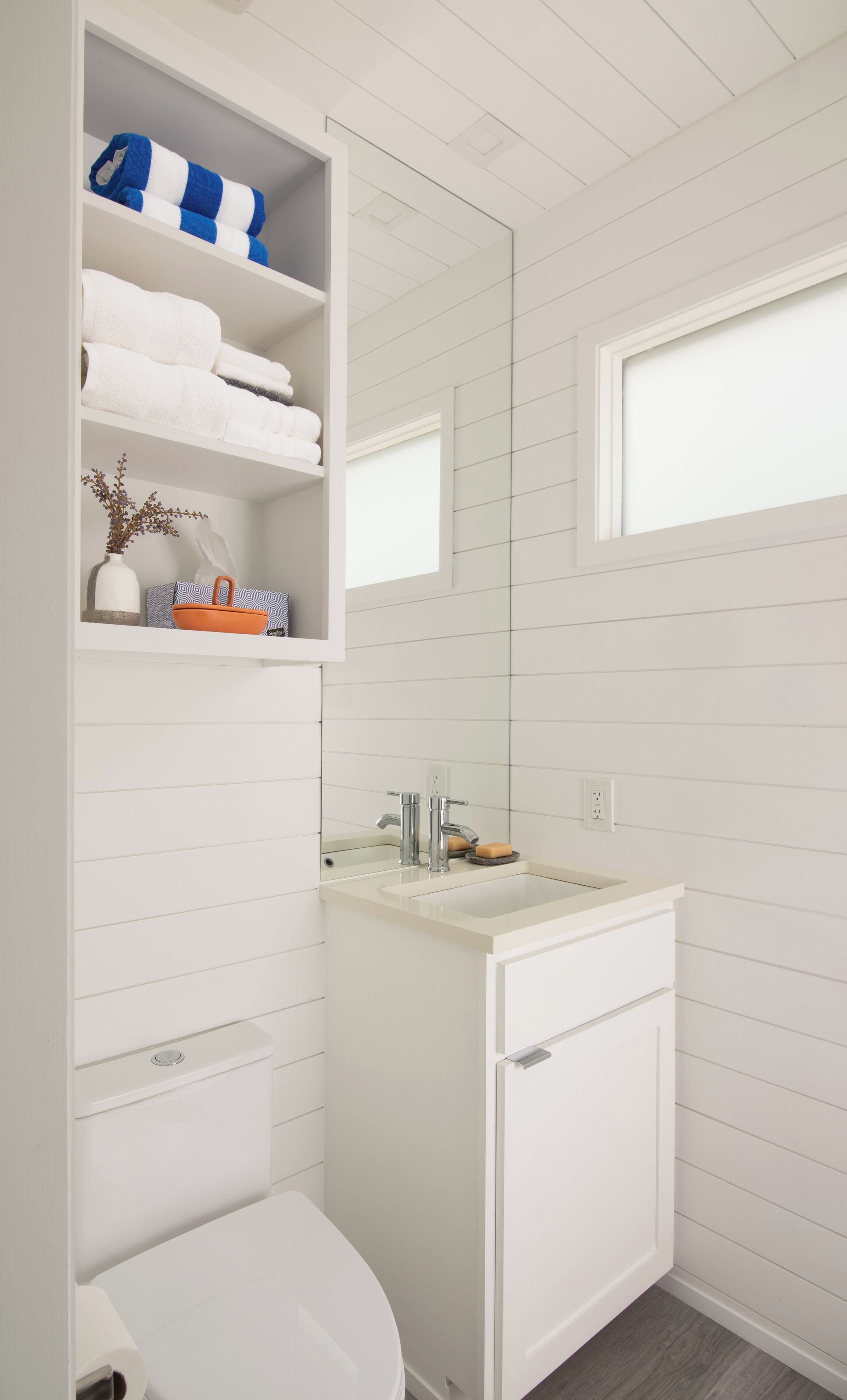 A bathroom with a white sink, mirror, and toilet. There are shelves with towels and toiletries.