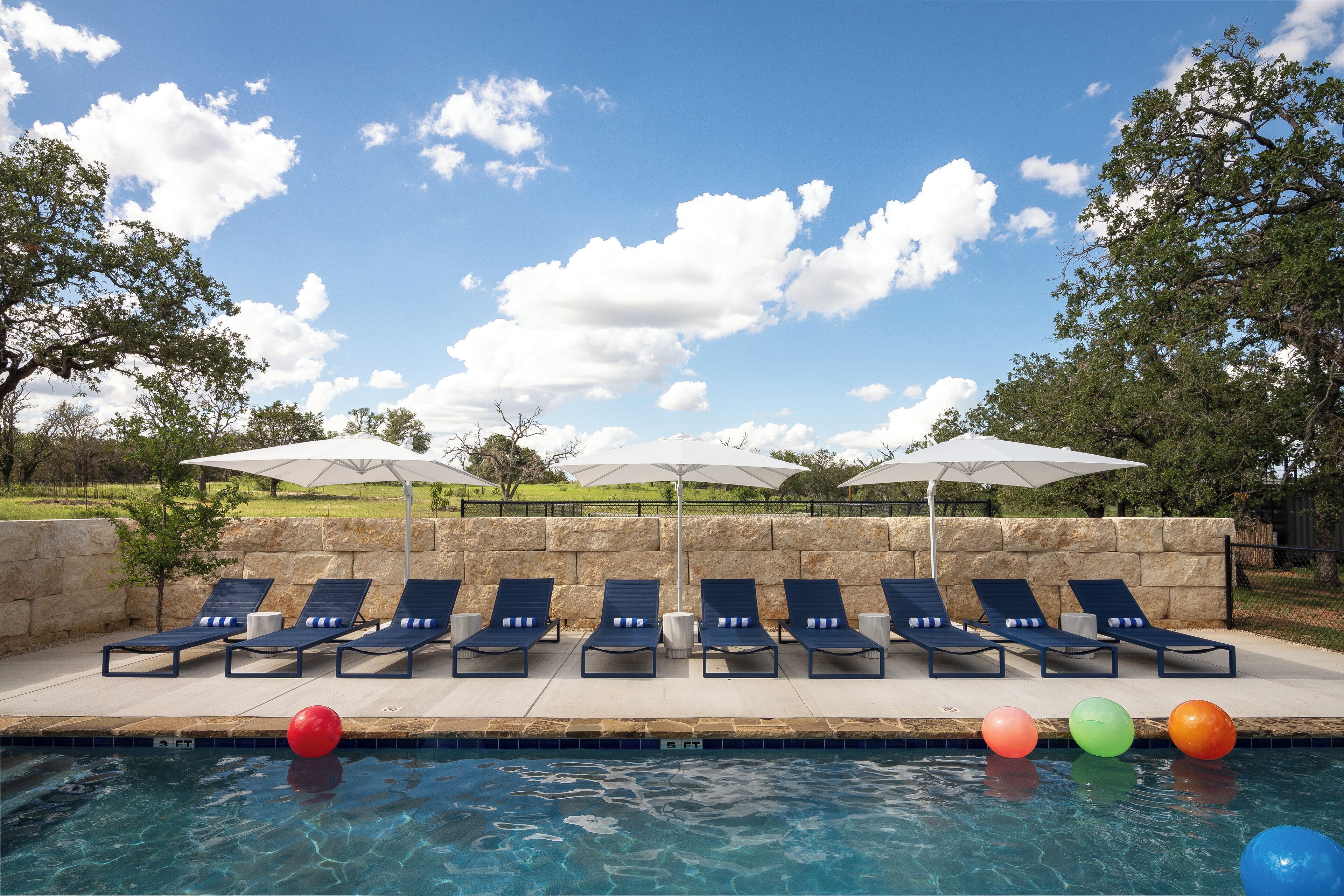 A poolside area with lounge chairs and umbrellas, a clear sky, and a stone wall in the background.